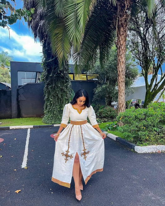 Woman in a white dress with gold accents standing in a parking lot with trees in the background. Simple Golden Zuria Habesha Kemis New Elegant Ethiopian Dress ethgebya gebeya