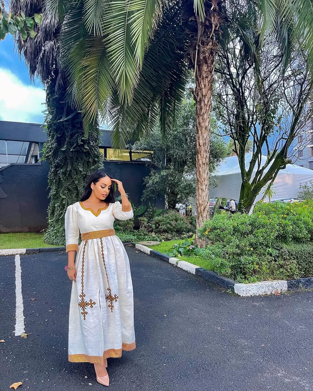 Woman in a white dress with gold accents standing on a road with trees and buildings in the background Simple Golden Zuria Habesha Kemis New Elegant Ethiopian Dress ethgebya gebeya