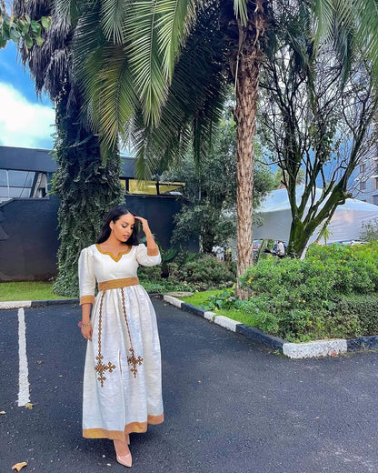 Woman in a white dress with gold accents standing on a road with trees and buildings in the background Simple Golden Zuria Habesha Kemis New Elegant Ethiopian Dress ethgebya gebeya