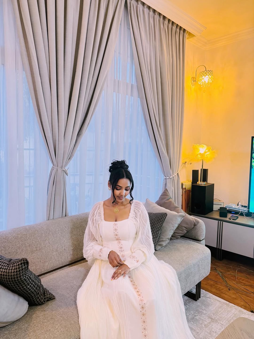 Woman in a white dress sitting on a couch in a living room with large windows and decorative pillows.  Long Sleeve Habesha Kemis | New Minimalist Ethiopian Traditional Dress ethgebya