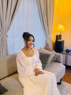 Woman in a white dress sitting on a couch in a living room with curtains and a lamp in the background.  Long Sleeve Habesha Kemis | New Minimalist Ethiopian Traditional Dress ethgebya