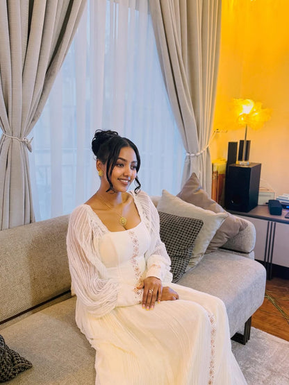 Woman in a white dress sitting on a couch in a living room with curtains and a lamp in the background.  Long Sleeve Habesha Kemis | New Minimalist Ethiopian Traditional Dress ethgebya