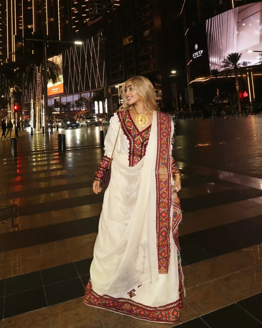 Woman in a traditional white and red embroidered outfit standing on a city street at night. Red Zuria Habesha Kemis | Handcrafted Ethiopian & Eritrean Fetil Dress Ethgebya