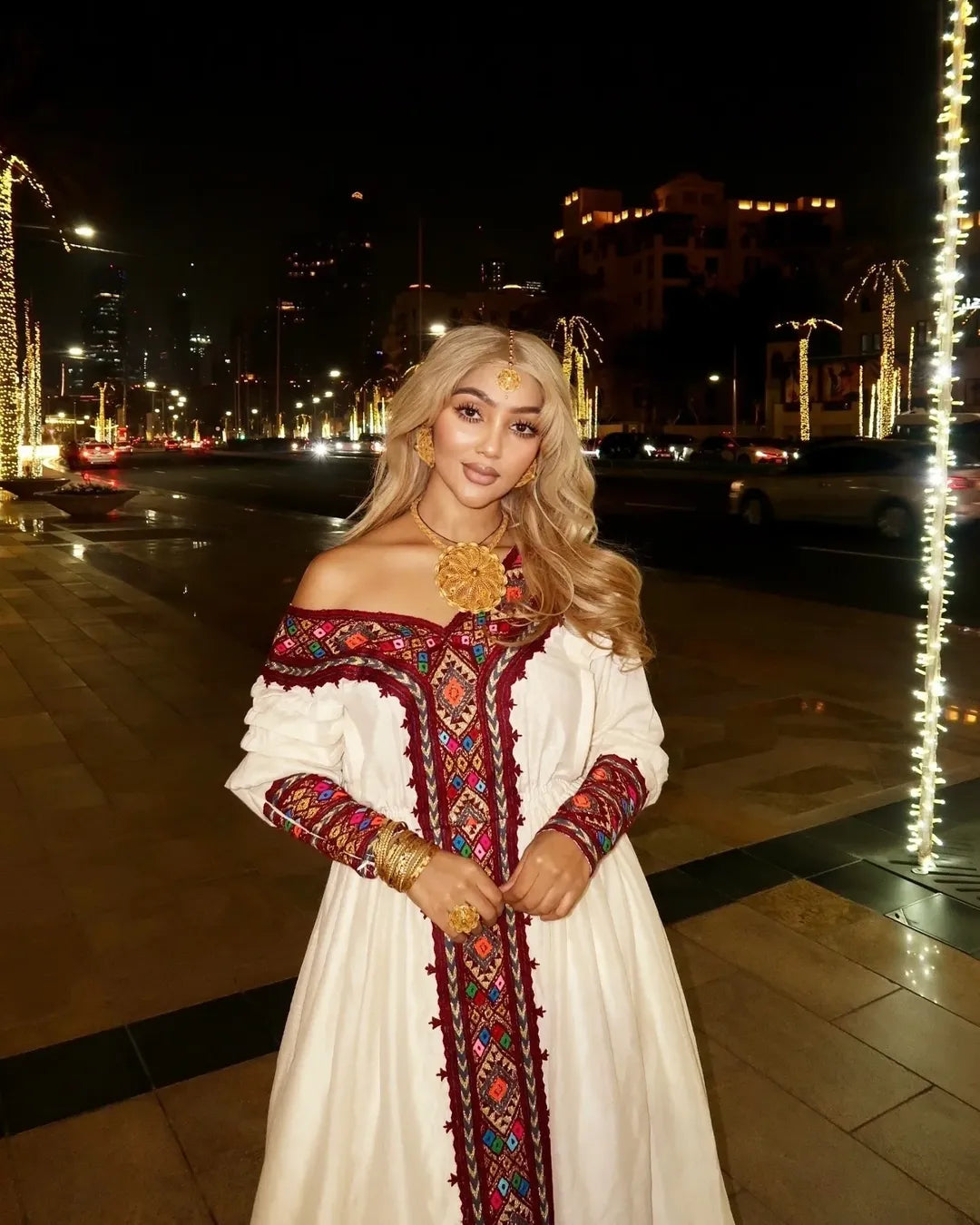 Woman in a traditional embroidered dress standing on a decorated street at night. Red Zuria Habesha Kemis | Handcrafted Ethiopian & Eritrean Fetil Dress Ethgebya