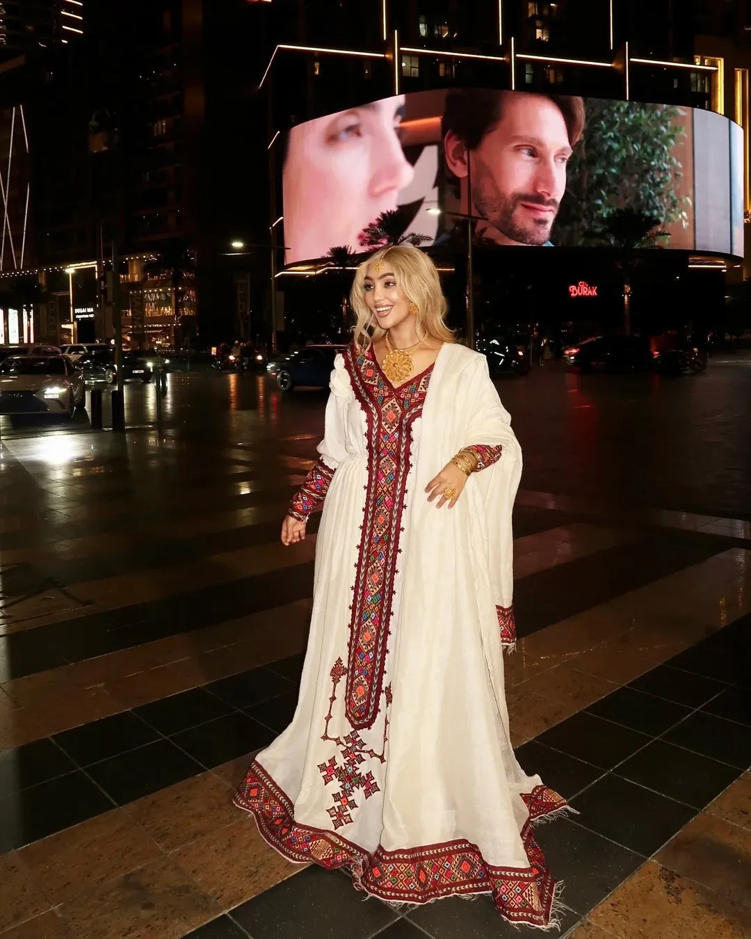 Woman in a white dress with red embroidery standing in front of a large screen displaying a movie scene. Red Zuria Habesha Kemis | Handcrafted Ethiopian & Eritrean Fetil Dress Ethgebya