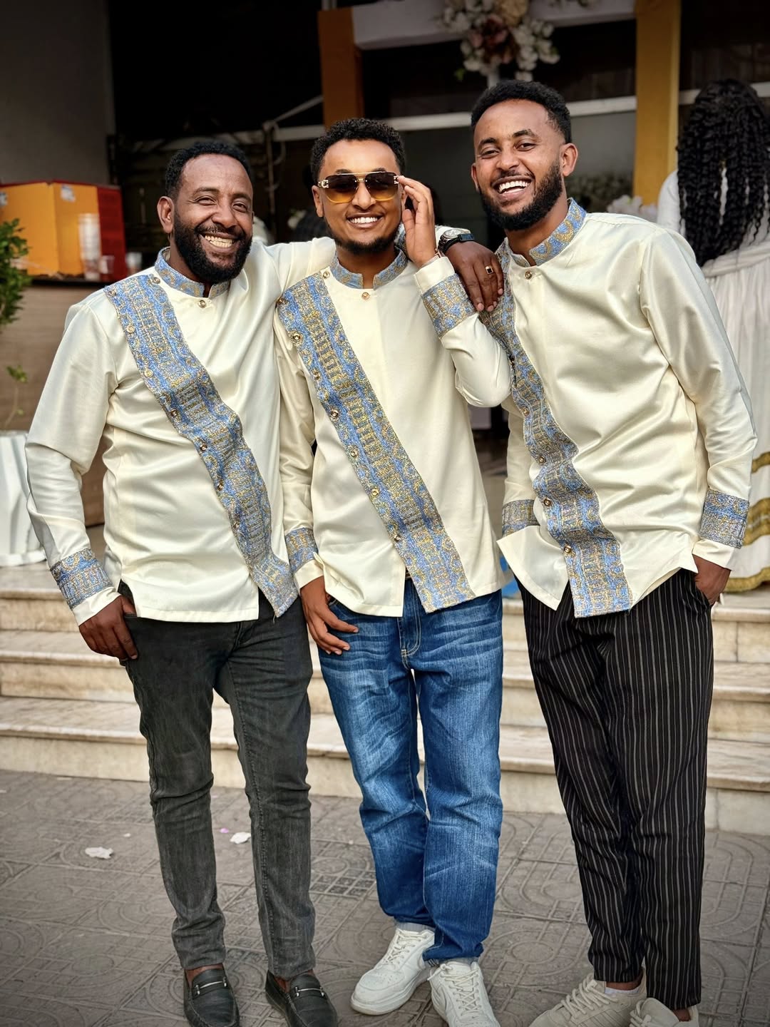 Three men wearing matching outfits with decorative sashes standing together outdoors. Modern Habesha Shirt for Men New Ethiopian Cultural Tilet Clothing Ethgebya