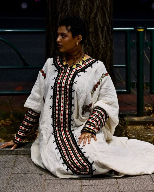 Woman wearing a white dress with black and red embroidery sitting on the ground. New Gondar Habesha Kemis Classic Ethiopian Traditional Dress Ethiopia Gebeya Ethgebya
