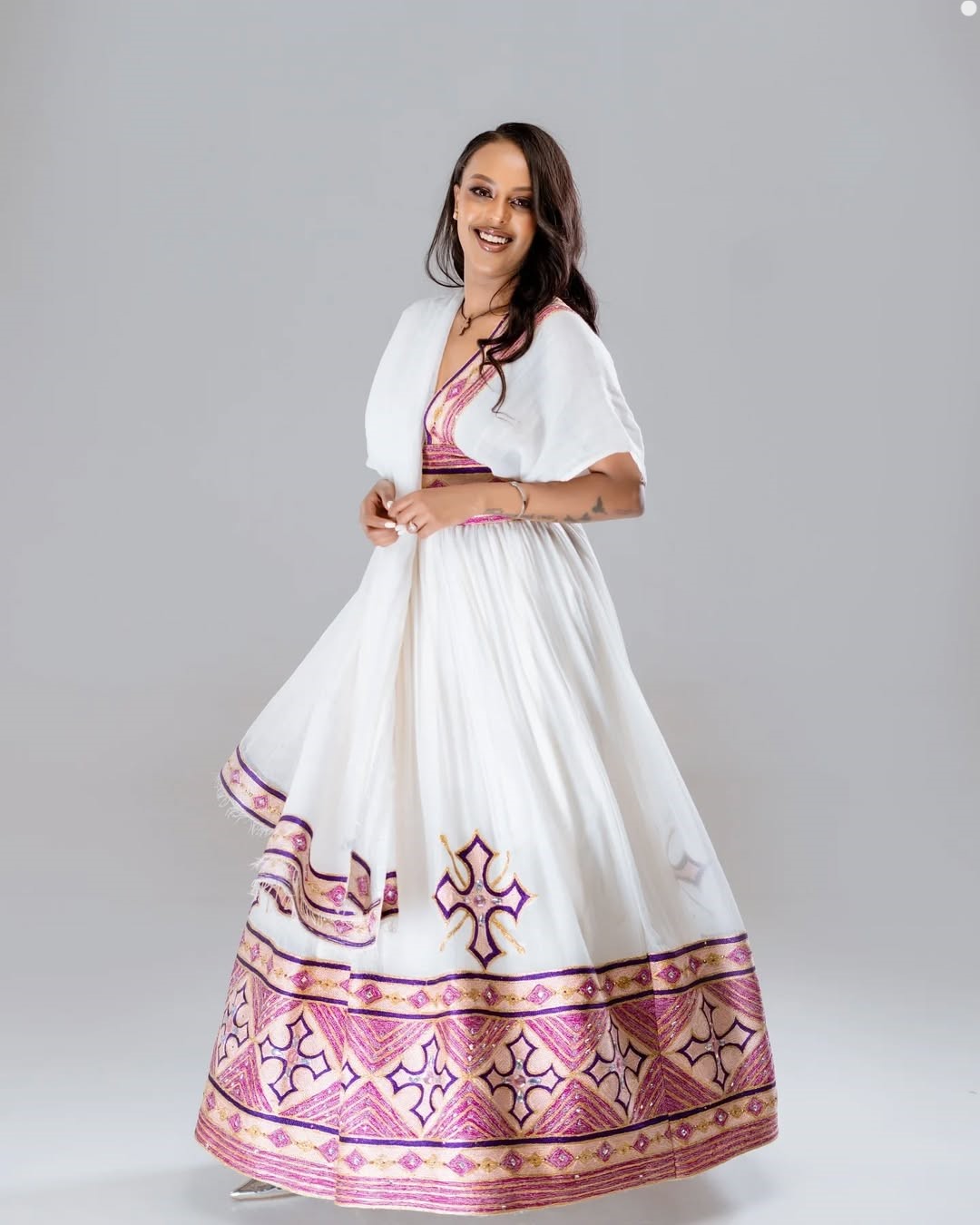 Woman wearing a white dress with pink and purple patterns on a gray background. Rose Gold Modern Zuria Habesha Kemis Handcrafted Ethiopian Dress ethgebya gebeya usa