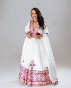 Woman wearing a white dress with pink and purple patterns on a plain background. Rose Gold Modern Zuria Habesha Kemis Handcrafted Ethiopian Dress ethgebya gebeya usa