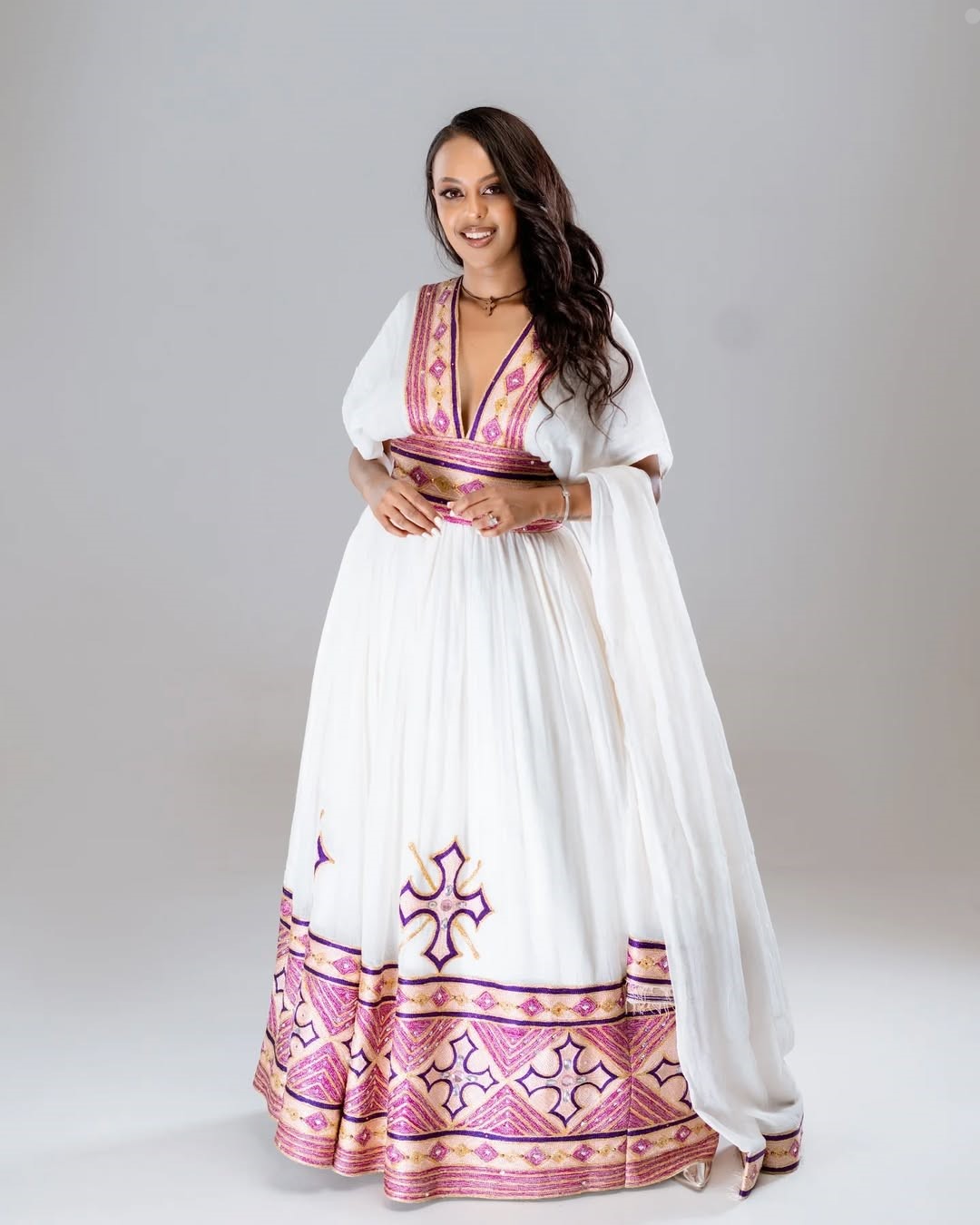 Woman wearing a white dress with pink and purple patterns on a plain background. Rose Gold Modern Zuria Habesha Kemis Handcrafted Ethiopian Dress ethgebya gebeya usa
