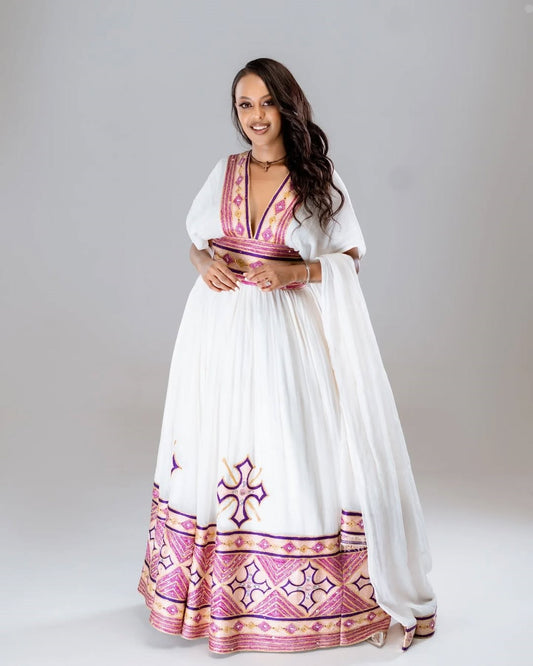 Woman wearing a white dress with pink and purple patterns on a plain background. Rose Gold Modern Zuria Habesha Kemis Handcrafted Ethiopian Dress ethgebya gebeya usa