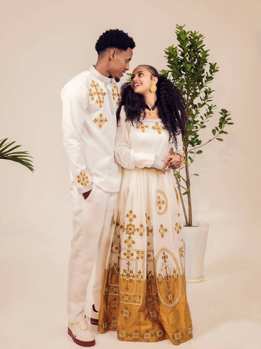Two people in traditional attire standing next to a plant against a plain background Ethiopian gown, Habesha wedding dress, Eritrean evening dress, Ethiopian traditional gown, Ethiopian heritage dress, Habesha chic, Eritrean cultural attire, Habesha elegant dress, Eritrean bridal wear, Ethiopian vintage dress, Habesha bridesmaid dress, African ethnic dress, Ethiopian handcrafted dress, Eritrean handcrafted dress, traditional African gown, Habesha festive dress, Ethiopian formal wear,