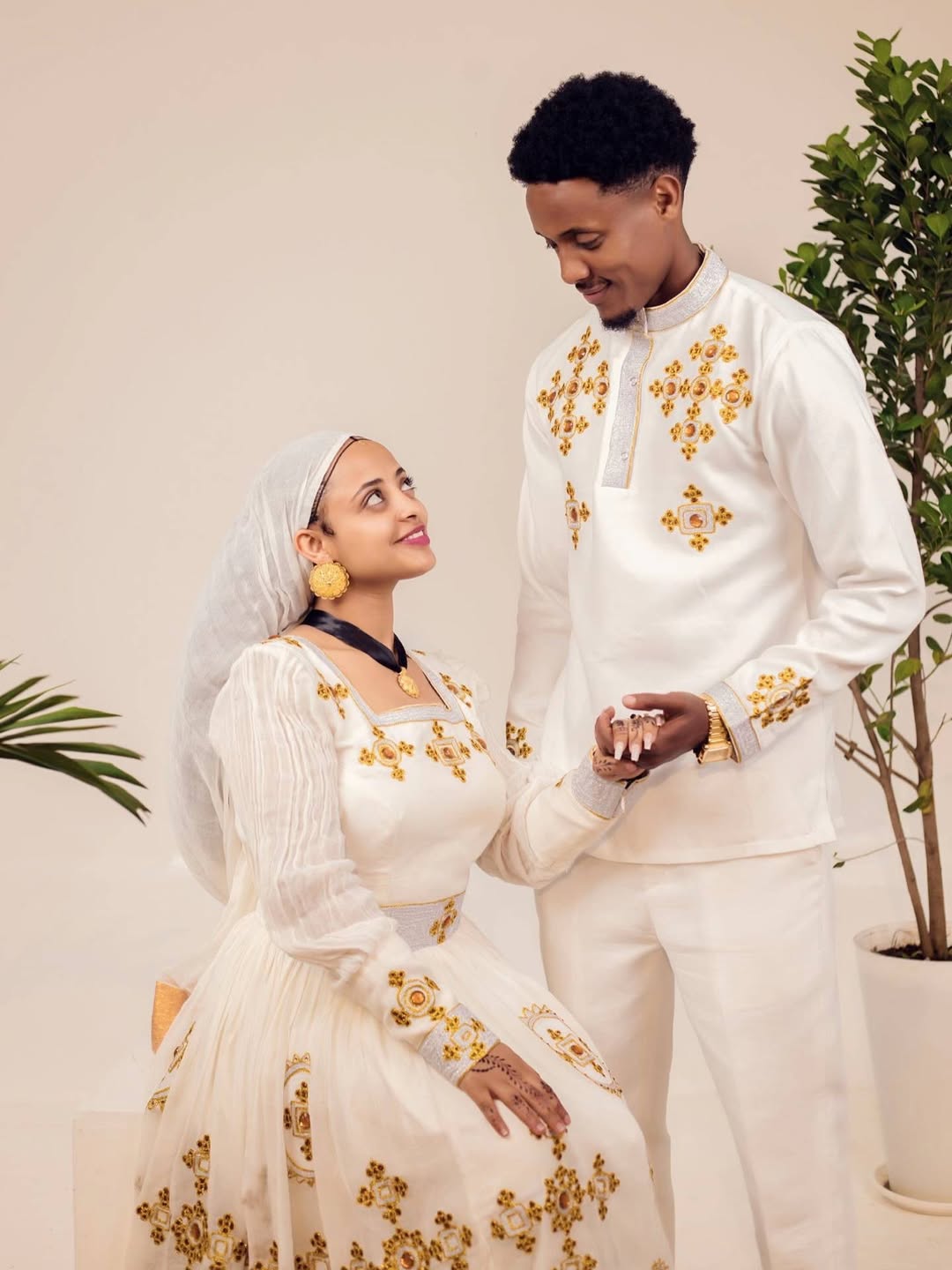 Man and woman in formal white outfits with gold embroidery standing close together. Ethiopian gown, Habesha wedding dress, Eritrean evening dress, Ethiopian traditional gown, Ethiopian heritage dress, Habesha chic, Eritrean cultural attire, Habesha elegant dress, Eritrean bridal wear, Ethiopian vintage dress, Habesha bridesmaid dress, African ethnic dress, Ethiopian handcrafted dress, Eritrean handcrafted dress, traditional African gown, Habesha festive dress, Ethiopian formal wear,