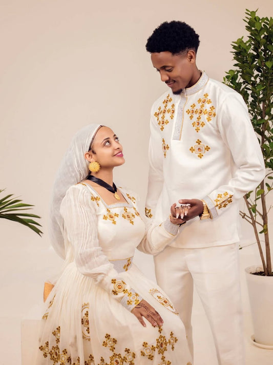 Man and woman in formal white outfits with gold embroidery standing close together. Ethiopian gown, Habesha wedding dress, Eritrean evening dress, Ethiopian traditional gown, Ethiopian heritage dress, Habesha chic, Eritrean cultural attire, Habesha elegant dress, Eritrean bridal wear, Ethiopian vintage dress, Habesha bridesmaid dress, African ethnic dress, Ethiopian handcrafted dress, Eritrean handcrafted dress, traditional African gown, Habesha festive dress, Ethiopian formal wear,