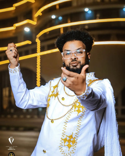 Man in a white outfit with gold embroidery pointing forward, with a blurred indoor background. Habesha inspired fashion, Ethiopian custom made dress, Habesha modern Kemis, bridal dress, wedding outfit, Ethiopian embroidered gown, ceremony dress, Habesha festive dress, Ethiopian cultural gown, elegant attire, Habesha handmade , heritage dress, festive gown, Eritrean festive outfit, lace Kemis, designer dress, artisan clothing, festive gown, premium , traditional cotton attire, embroidered gown, handmade clot