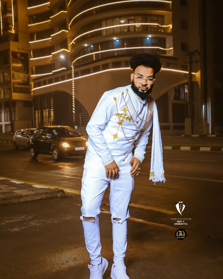 Man in a white outfit with gold accents standing on a street at night. Habesha inspired fashion, Ethiopian custom made dress, Habesha modern Kemis, bridal dress, wedding outfit, Ethiopian embroidered gown, ceremony dress, Habesha festive dress, Ethiopian cultural gown, elegant attire, Habesha handmade , heritage dress, festive gown, Eritrean festive outfit, lace Kemis, designer dress, artisan clothing, festive gown, premium , traditional cotton attire, embroidered gown, handmade clothing, festive clothing, 
