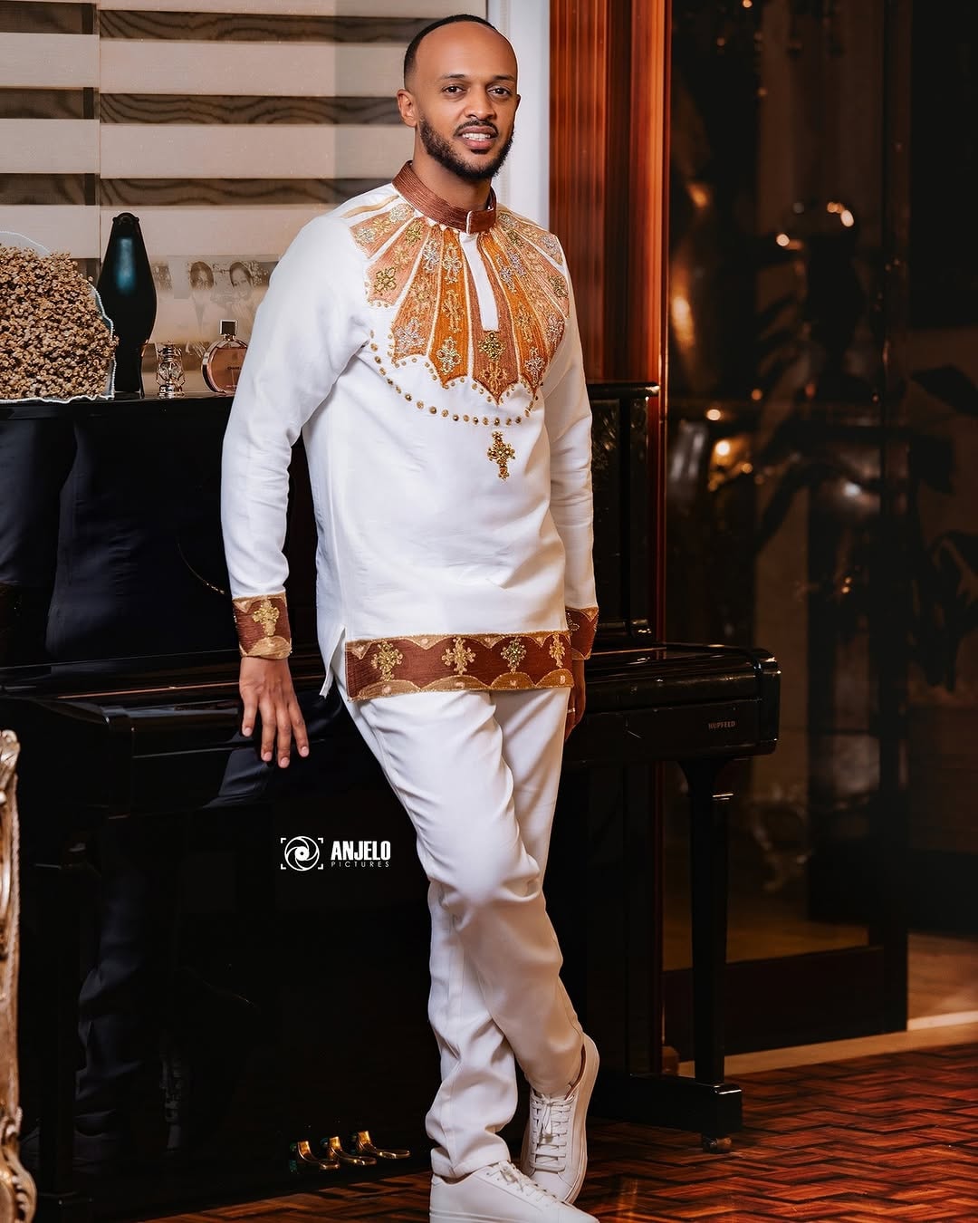Man in a white and gold traditional outfit standing in a room with decorative elements. Brown Habesha Men’s Shirt New Modern Ethiopian Traditional Wear ethgebya gebeya habesha men