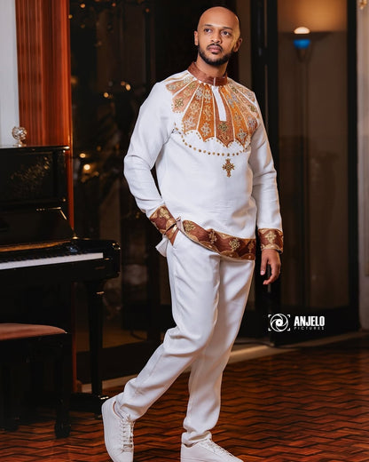 Man in a white outfit with intricate patterns standing in a room with a piano. Brown Habesha Men’s Shirt New Modern Ethiopian Traditional Wear ethgebya gebeya habesha men