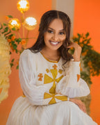 Woman in a white and gold dress sitting against an orange background with decorative elements. Golden Ethiopian Habesha Dress Handcrafted Zuria Kemis Set ethgebya gebeya usa zuria habesha kemis 