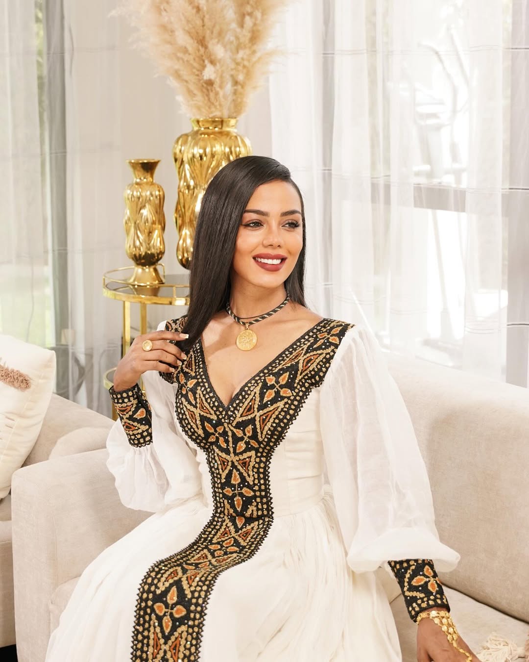 Woman in a white and black dress with gold accents sitting on a couch in a stylish room. Black & Gold Tilet Zuria Habesha Kemis New Modern Ethiopian Dress ethgebya gebeya habesha kemis zuria kemis