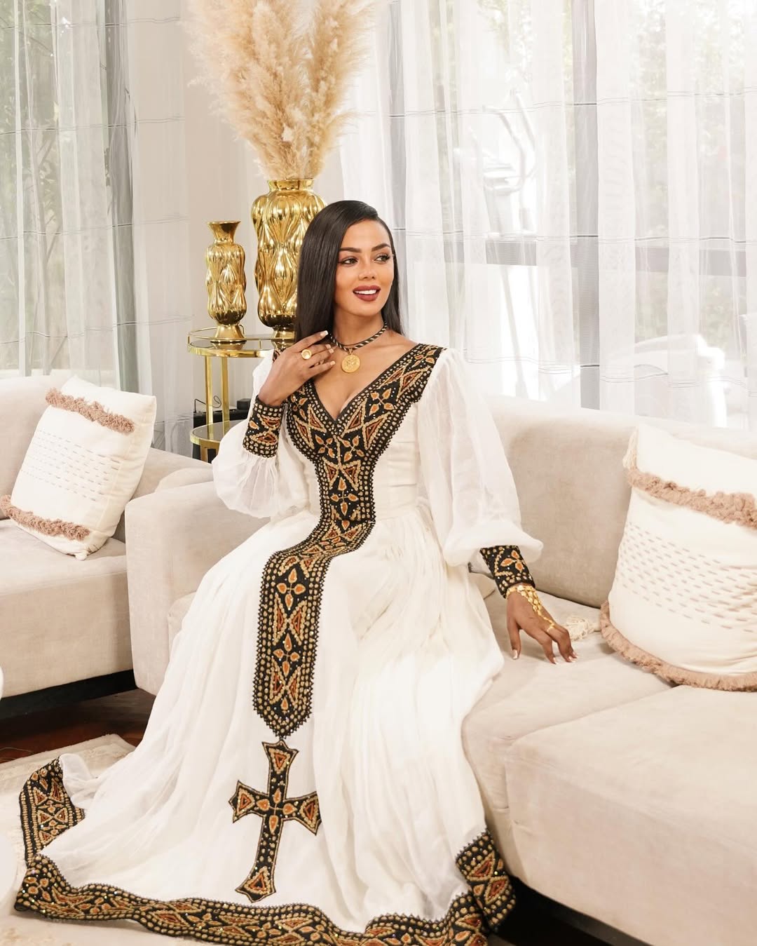 Woman in a white and black embroidered dress sitting on a couch in a stylish room. Black & Gold Tilet Zuria Habesha Kemis New Modern Ethiopian Dress ethgebya gebeya habesha kemis zuria kemis