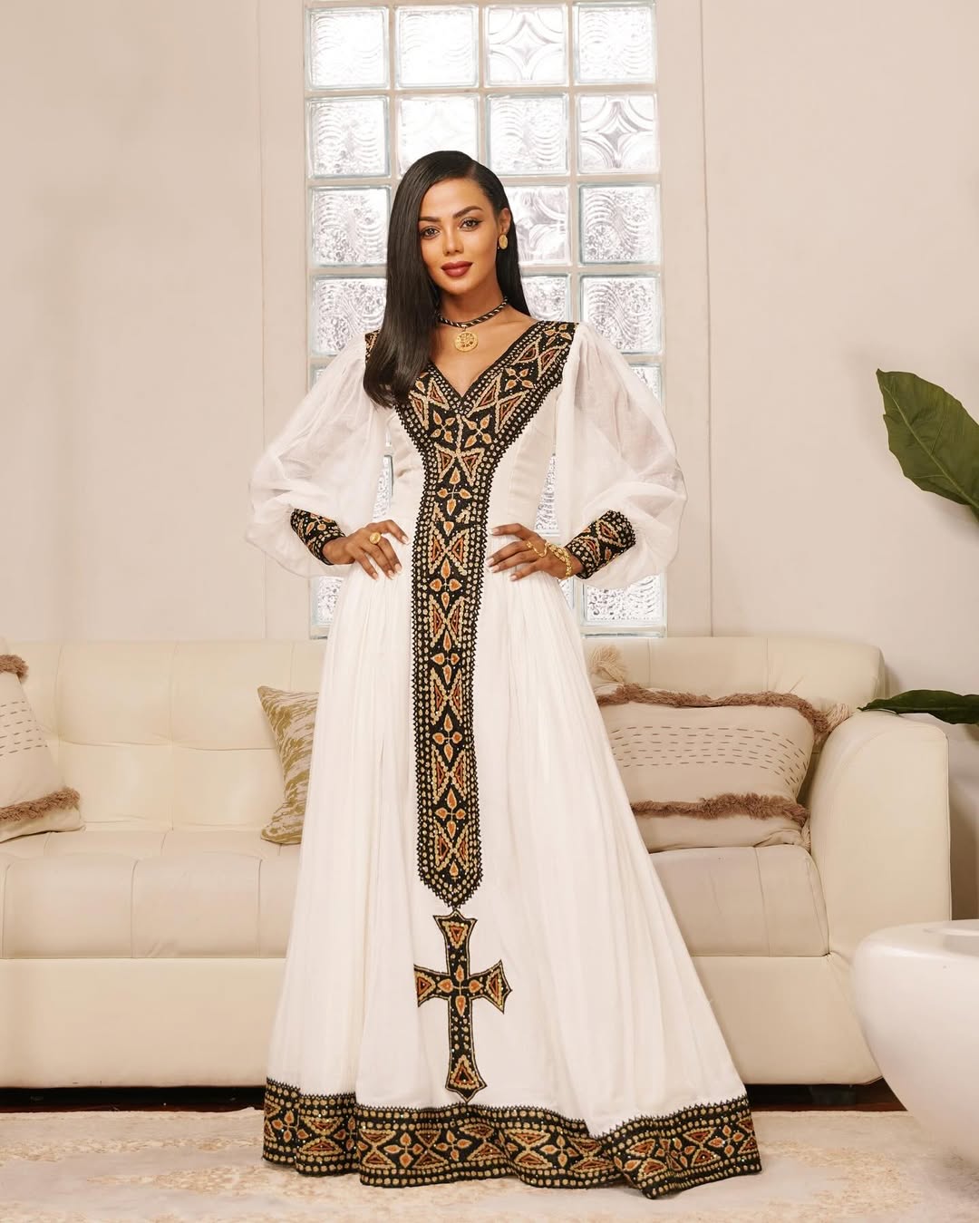 Woman wearing a white dress with black embroidery in a living room setting. Black & Gold Tilet Zuria Habesha Kemis New Modern Ethiopian Dress ethgebya gebeya habesha kemis zuria kemis