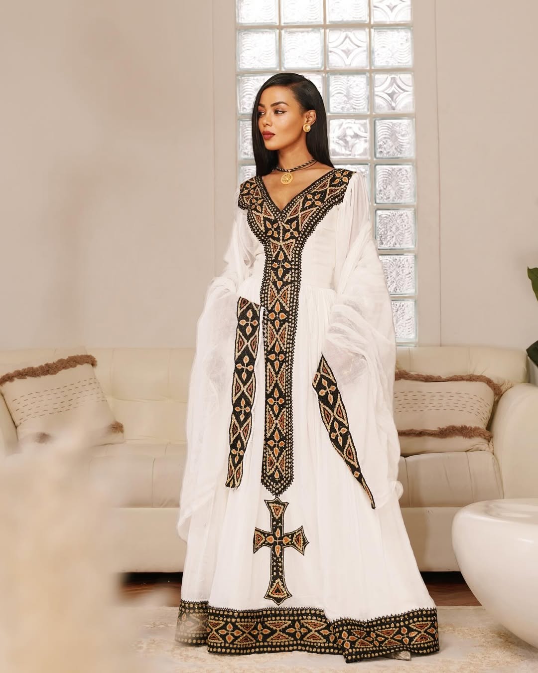 Woman wearing a white dress with black embroidery in a living room setting. Black & Gold Tilet Zuria Habesha Kemis New Modern Ethiopian Dress ethgebya gebeya habesha kemis zuria kemis