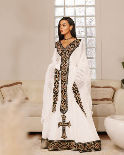 Woman wearing a white dress with black embroidery in a living room setting. Black & Gold Tilet Zuria Habesha Kemis New Modern Ethiopian Dress ethgebya gebeya habesha kemis zuria kemis