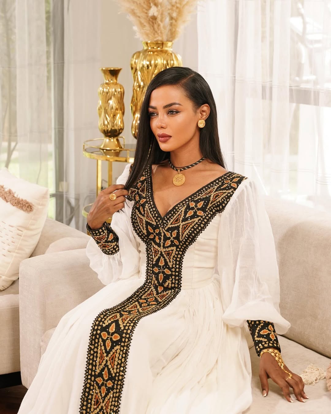 Woman in a white dress with black embroidery sitting on a couch in a stylish room. Black & Gold Tilet Zuria Habesha Kemis New Modern Ethiopian Dress ethgebya gebeya habesha kemis zuria kemis