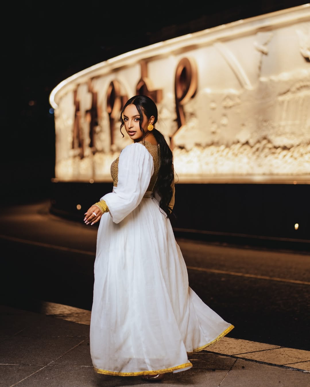Woman in a white dress standing in front of a large stone sculpture at night. Gold Zuria Habesha Kemis New Elegant Modern Ethiopian Dress ethgebya gebeya habesha kemis