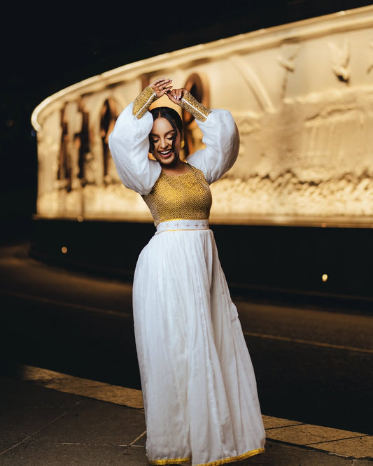 Woman in gold top and white skirt posing in front of an ancient Egyptian temple. Gold Zuria Habesha Kemis New Elegant Modern Ethiopian Dress ethgebya gebeya habesha kemis