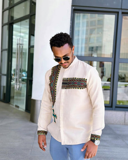 Man wearing a white embroidered shirt with colorful patterns in an urban setting Cream Luxury Habesha Men’s Shirt New Modern Ethiopian Style ethgebya gebeya wen wond habehsa