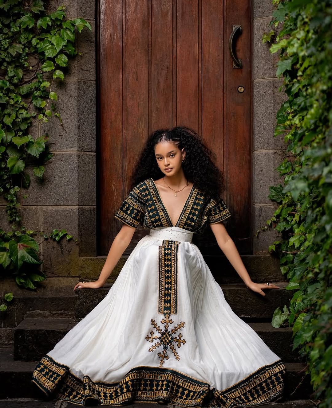 Woman in a white dress with black and gold embroidery sitting on steps outdoors. Golden & Green Modern Habesha Kemis Elegant Ethiopian ethgebya gebeyaDress