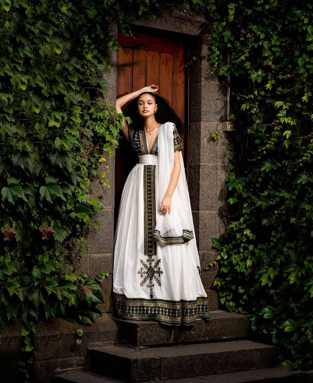 Woman in a white and black traditional outfit standing in a doorway surrounded by greenery. Golden & Green Modern Habesha Kemis Elegant Ethiopian ethgebya gebeyaDress