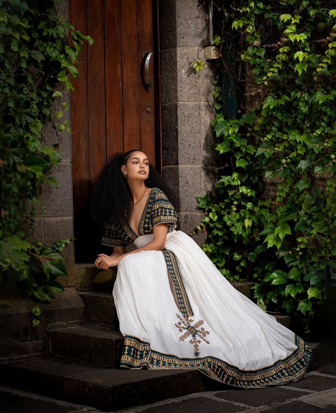 Woman in a white and black embroidered traditional outfit sitting on steps with greenery around. Golden & Green Modern Habesha Kemis Elegant Ethiopian ethgebya gebeyaDress
