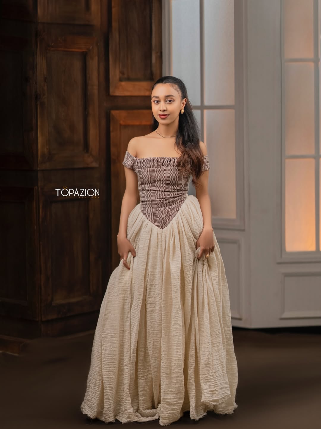 Woman wearing a patterned off-shoulder top and beige skirt with 'Topazion' branding. Modern Brown & Beige Habesha Kemis ethgebya