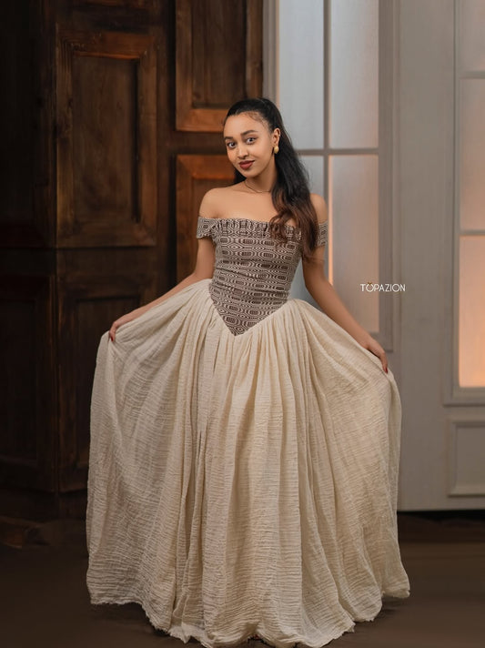 Woman in a patterned off-shoulder top and beige skirt standing indoors. Modern Brown & Beige Habesha Kemis ethgebya