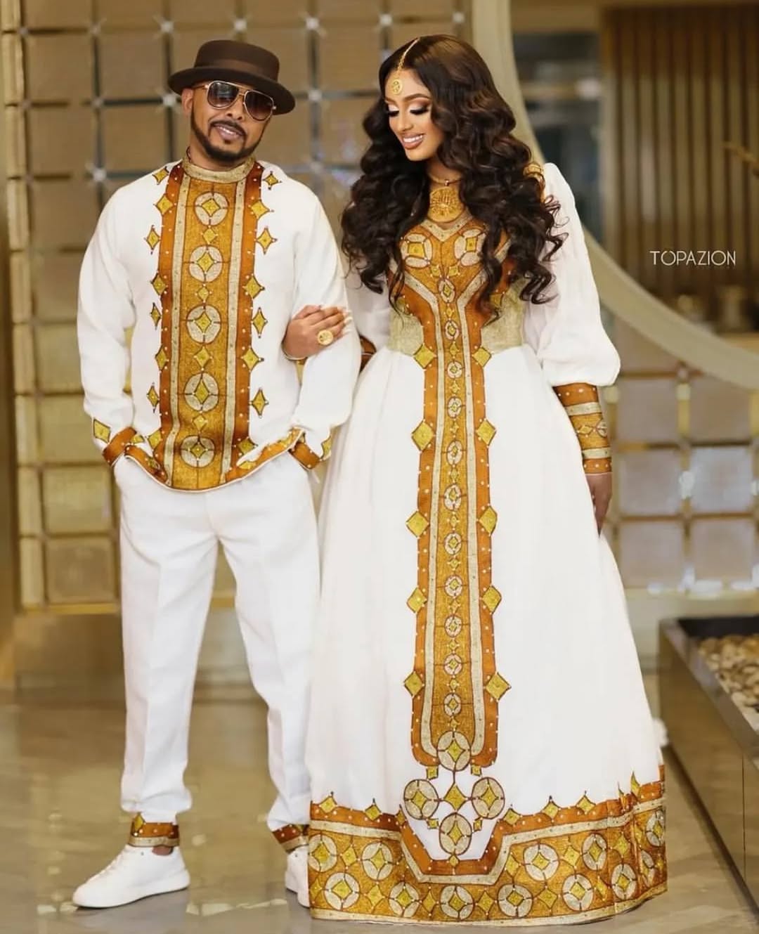 Man and woman in matching traditional outfits with gold embroidery in a decorative setting. Golden Ethiopian Habesha Couple Outfit | New Matching Modern Set