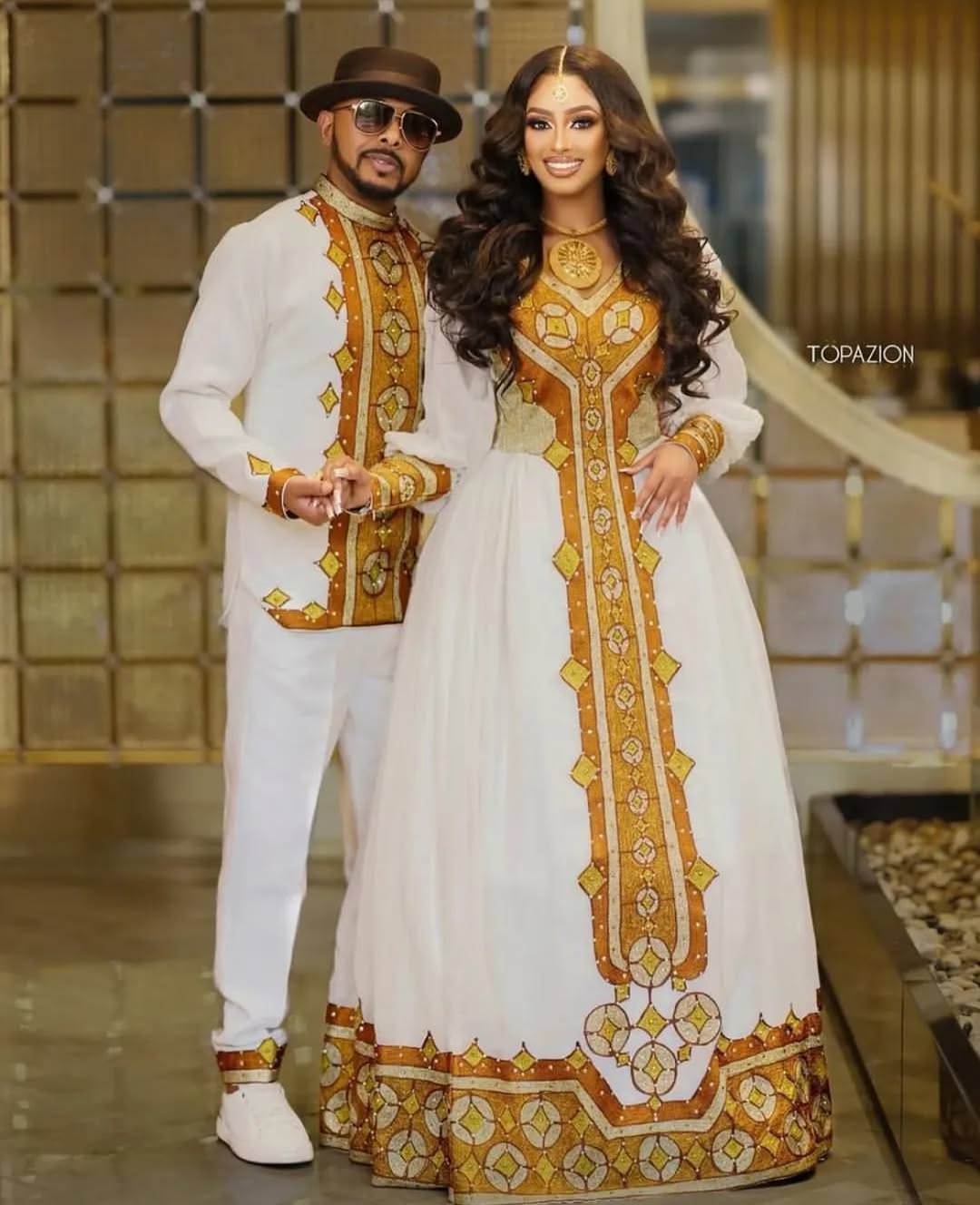 Man and woman in matching traditional outfits with gold and white patterns, standing indoors. Golden Ethiopian Habesha Couple Outfit | New Matching Modern Set