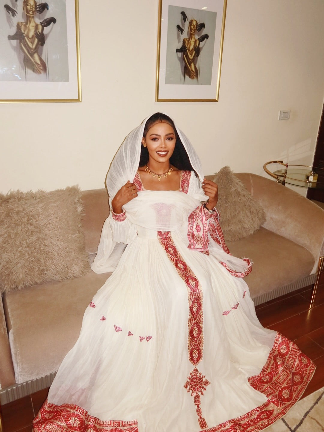 Woman in a white and red embroidered dress sitting on a couch with framed artwork in the background. New Pink Zuria Habesha Kemis Handcrafted Ethiopian Traditional Dress Ethiopia Gebebya Ethgebya