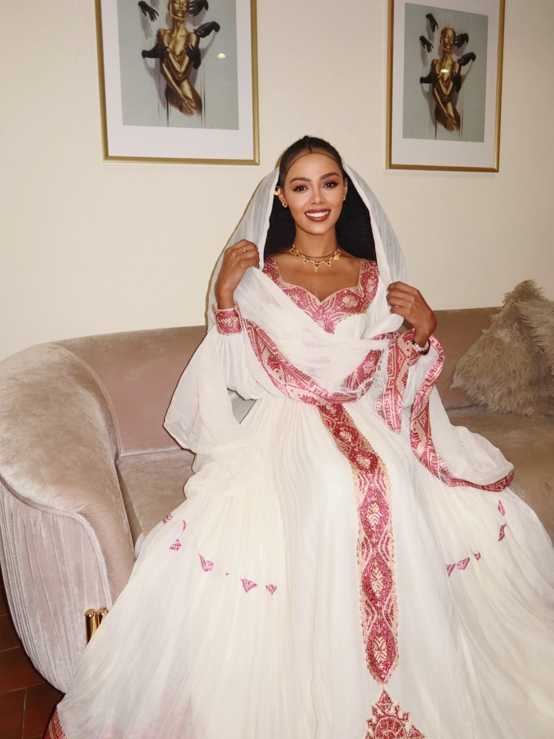 Woman in a white and red embroidered dress sitting on a couch with framed artwork in the background. New Pink Zuria Habesha Kemis Handcrafted Ethiopian Traditional Dress Ethiopia Gebebya Ethgebya