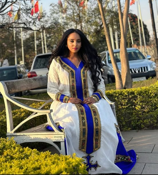 Woman in a white and blue traditional outfit sitting on a bench outdoors.  Elegant Handmade Habesha Kemis | Ethiopian Traditional Dress for Women ethgebya gebeya usa