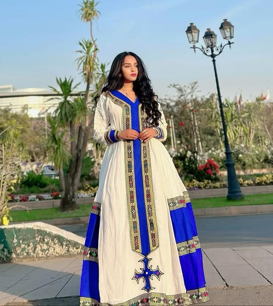 Woman in a white and blue traditional outfit with intricate designs standing outdoors.  Elegant Handmade Habesha Kemis | Ethiopian Traditional Dress for Women ethgebya gebeya usa