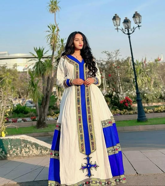 Woman in a white and blue traditional outfit with intricate designs standing outdoors.  Elegant Handmade Habesha Kemis | Ethiopian Traditional Dress for Women ethgebya gebeya usa