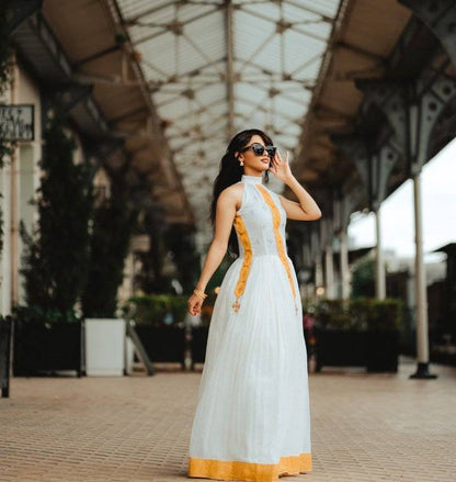 Woman in a white and orange dress standing in an outdoor setting with a geometric roof structure. Yellow Modern Zuria Habesha Kemis Handcrafted Ethiopian Dress ethgebya gebeya usa habesha kemis