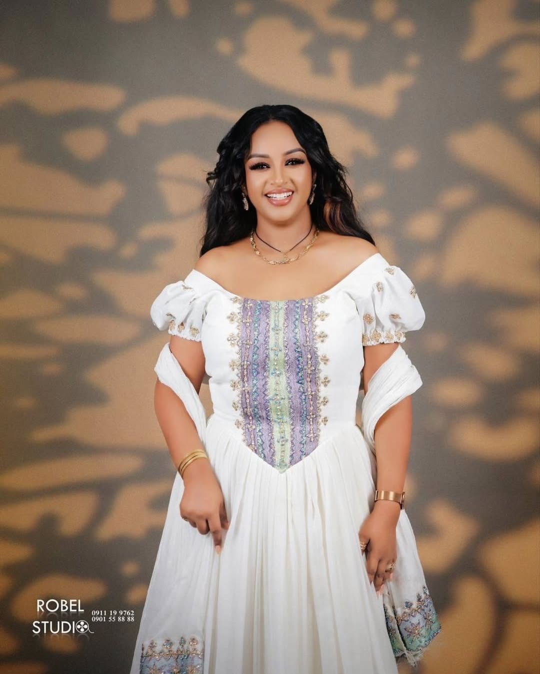 Woman in a white dress with colorful patterns against a patterned background. Modern Luxury Zuria Kemis Elegant Ethiopian Traditional Dress ethgebya gebeya
