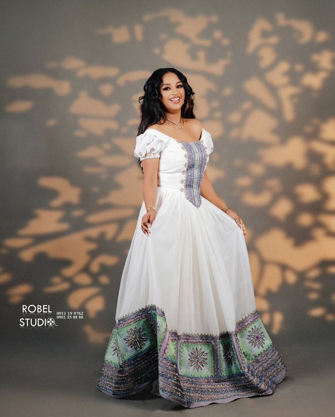 Woman in a white dress with patterned hem standing against a textured wall. Modern Luxury Zuria Kemis Elegant Ethiopian Traditional Dress ethgebya gebeya