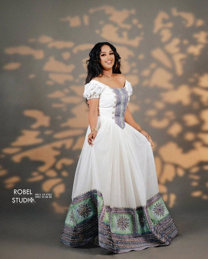Woman in a white dress with patterned hem standing against a textured wall. Modern Luxury Zuria Kemis Elegant Ethiopian Traditional Dress ethgebya gebeya
