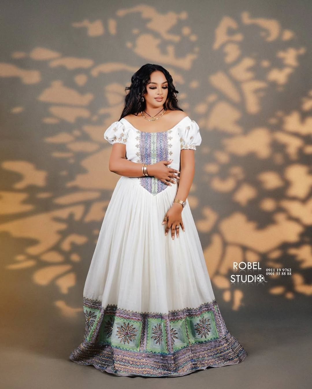 Woman in a white and patterned dress standing against a textured wall with 'Robel Studio' branding. Modern Luxury Zuria Kemis Elegant Ethiopian Traditional Dress ethgebya gebeya