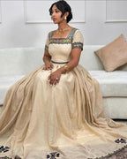 Woman in a beige saree with intricate designs sitting on a couch. New Beige Zuria Habesha Kemis Modern Handcrafted Ethiopian Dress ethgebya gebeya